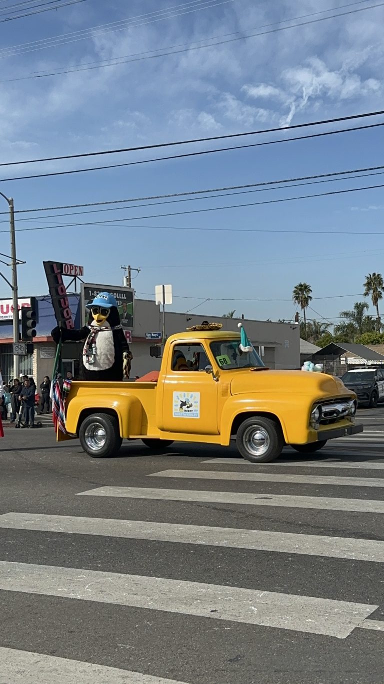 A wide shot of a yellow truck owned by Norky, Built For Kids TV. A green Santa hat is on the windshield. A sombrero is on the roof of the truck. Standing on the trunk of the truck is the penguin mascot, wearing a scarf and blue baseball cap.