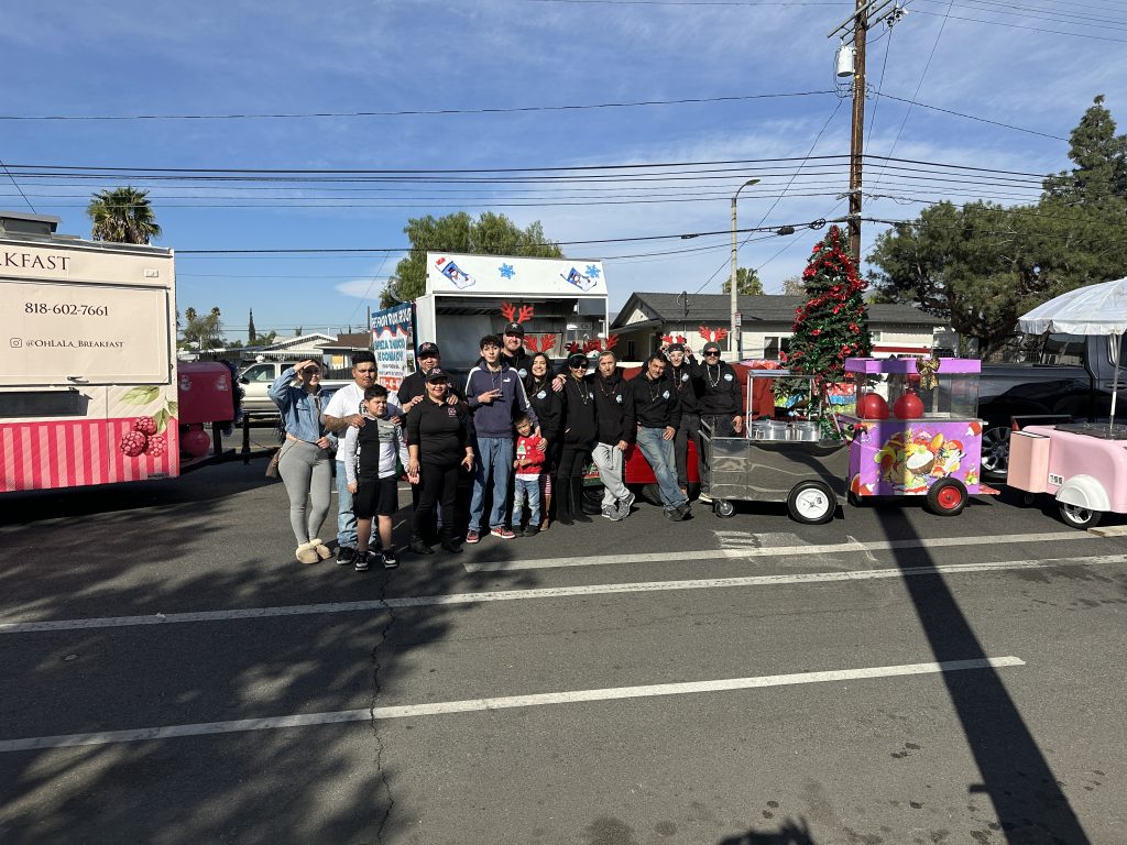 A wide shot of The Food Truck Group and Oh La-La Coffee and Breakfast families and guests, They stand in a row for a picture. In the center is a red sofa, where some of the people sit. Beside them on the right are two food carts. To the left is the back of Oh La-La's food truck.