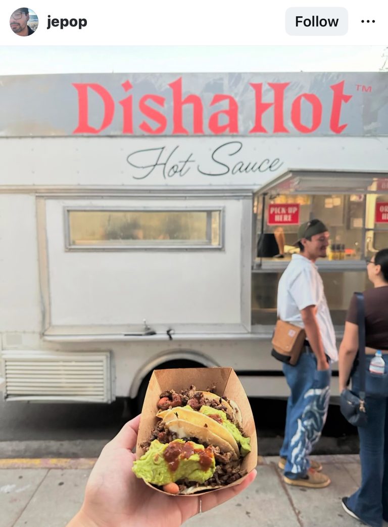 Wide shot of Disha Hot food truck. Customers wait in line. In front of the camera is a hand holding tacos covered in Disha Hot sauce.