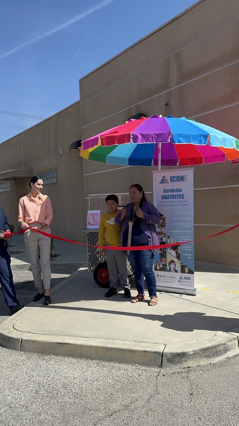 Wide shot of Yudy Mena holding the ribbon cutting scissors. She stands beside her son. ICON CDC guests hold the ribbon for Yudy. Behind Yudy is her fruit cart, roll-up banner, and rainbow umbrella.