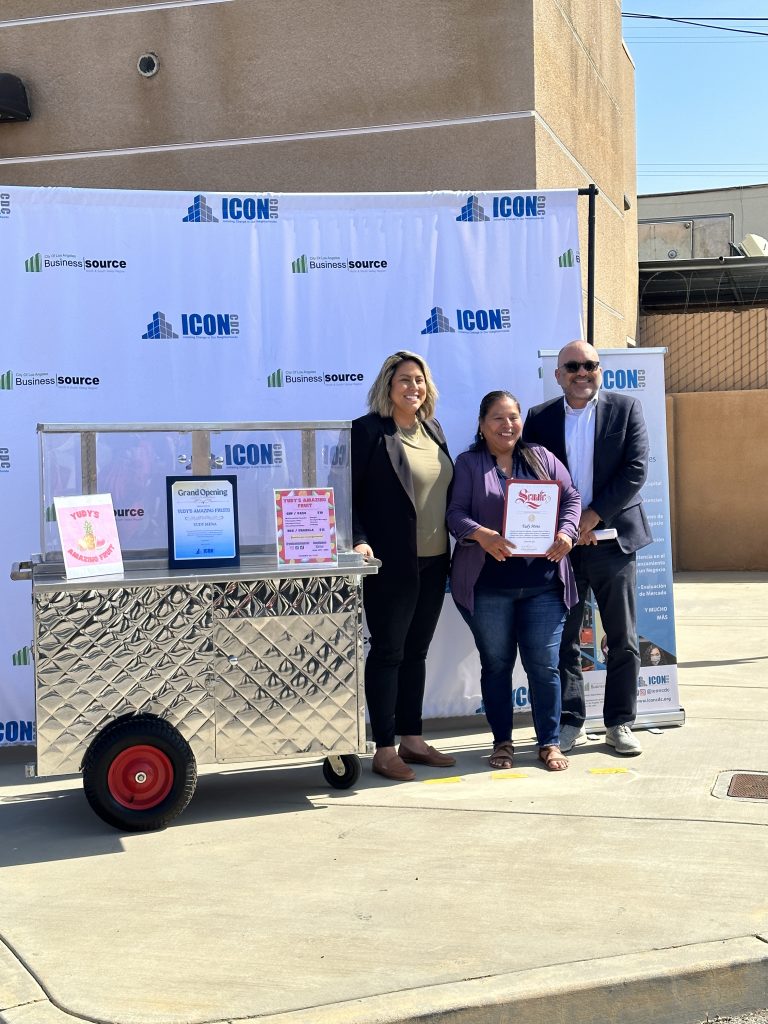 A wide shot of Yudy Mena holding her reward. Senator Caroline Menjivar and an ICON CDC representative stand on either side of her. Beside them is Rudy's fruit cart. Behind them is an ICON CDC's backdrop and roll-up banner.