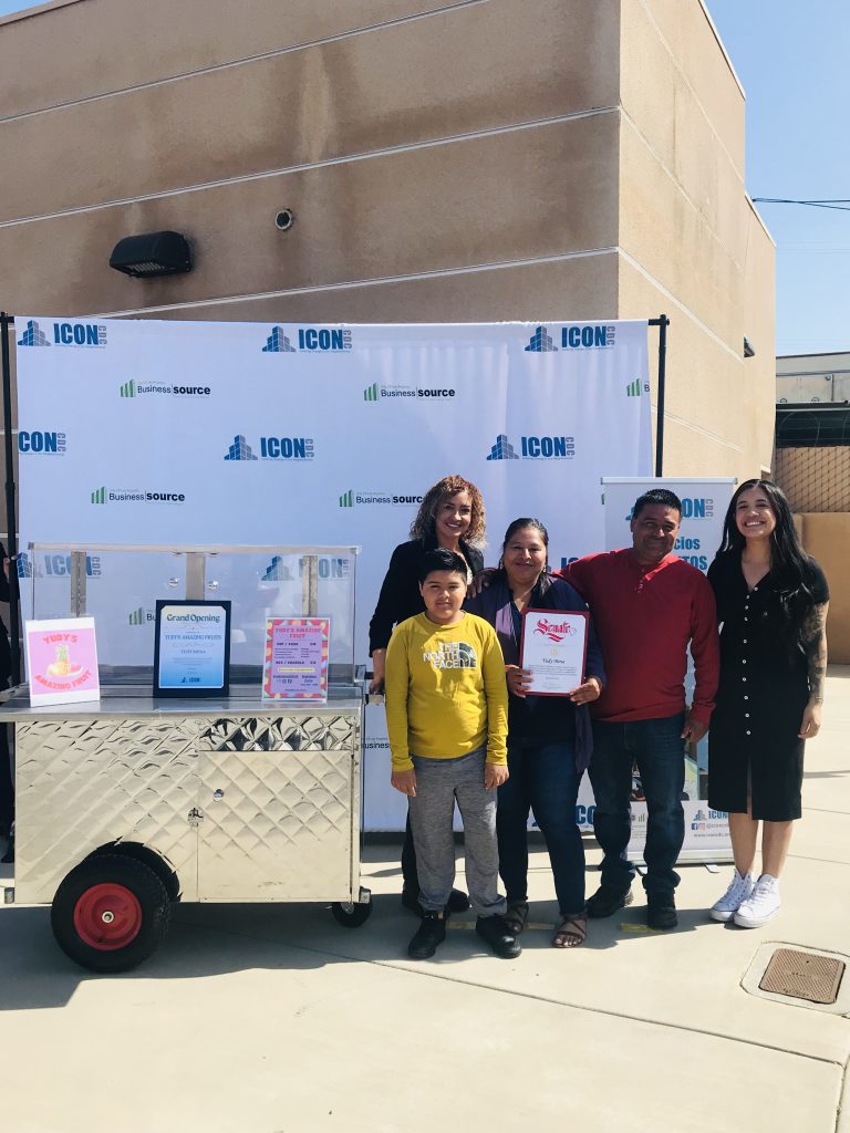 Wide shot of Yudy Mena holding her award. Yudy's family surrounds her. The Food Truck Group's owners, Maria Ponce and Kimberly Tapia, stand on either side. Beside them is Rudy's fruit card. Behind them is the ICON CDC backdrop and ICON CDC roll-up banner.