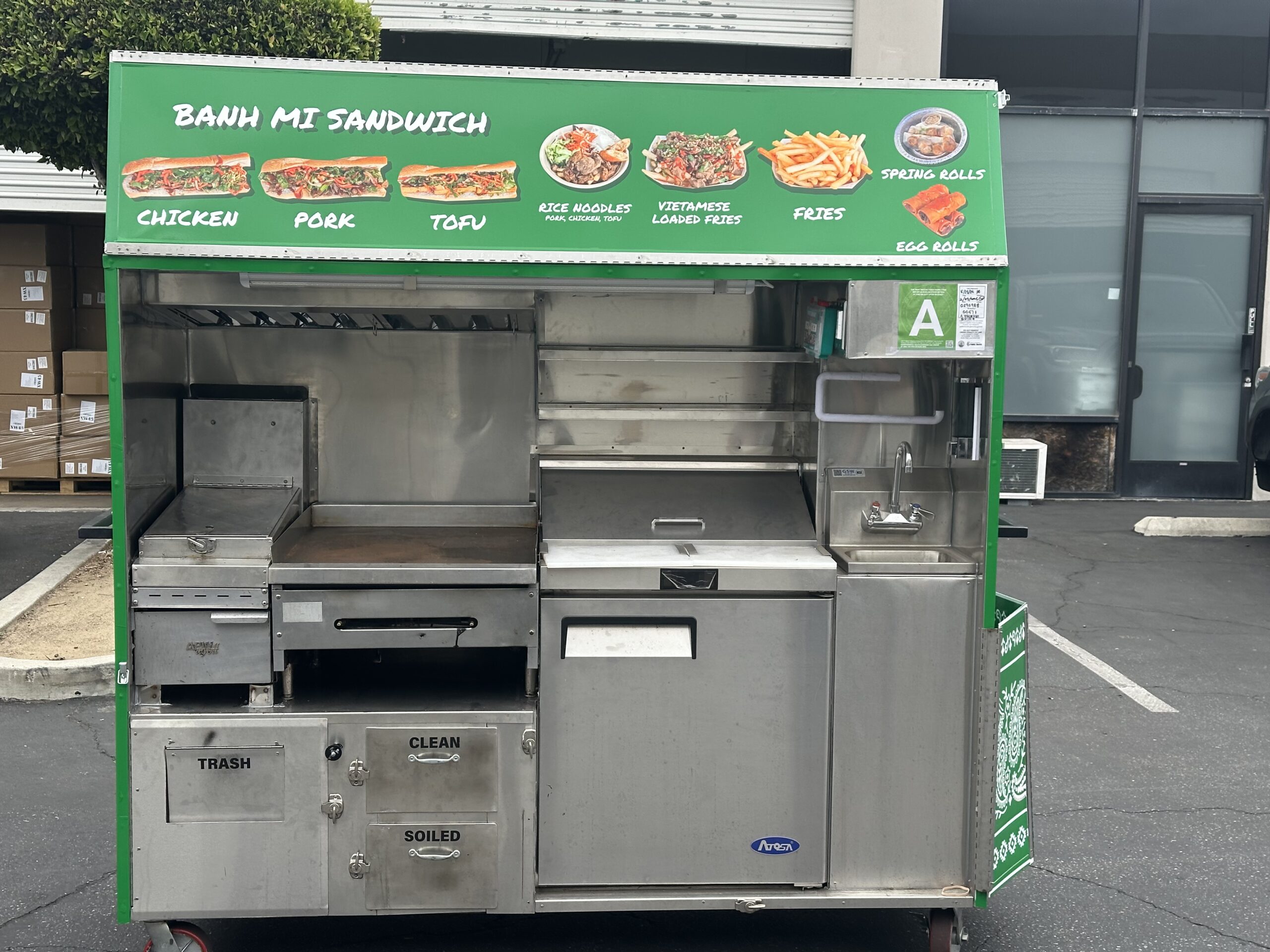A wide shot of a green food cart for Bang Mi Grill. The side is open to reveal the equipment view, including a sink and a prep table. The top of the food cart has a sandwich menu. The food cart is in a parking lot.