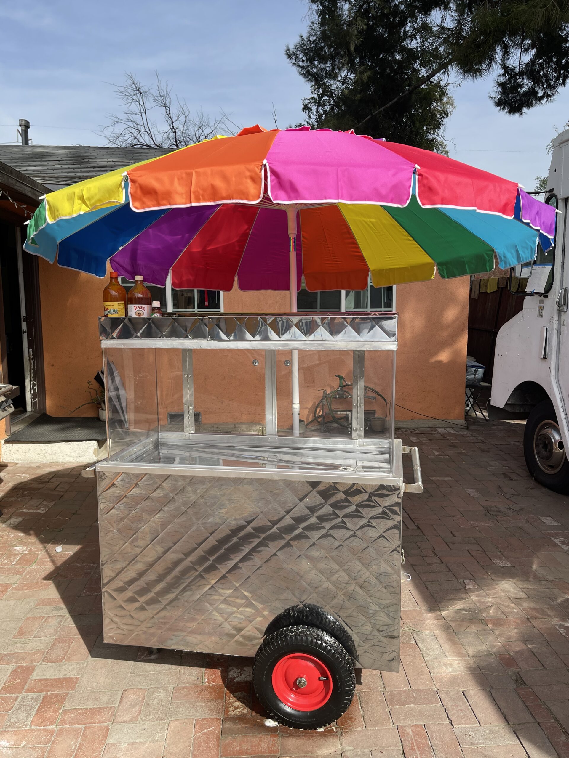 A wide shot of a fruit cart. The cart is made of steel. It has a glass view just above to look inside. Atop the food cart is a rainbow umbrella. The fruit cart is outside on a cloudy day.