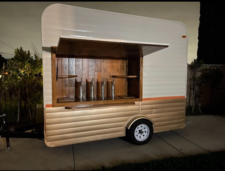 A wide shot of a beer tap trailer facing slightly sideways. The concession window is open. The interior features four taps. To the sides are small, wooden shelves. The interior is entirely made of wooden planks. The exterior of the trailer is white, with a brown lower half. Between the colors is a thin orange border.