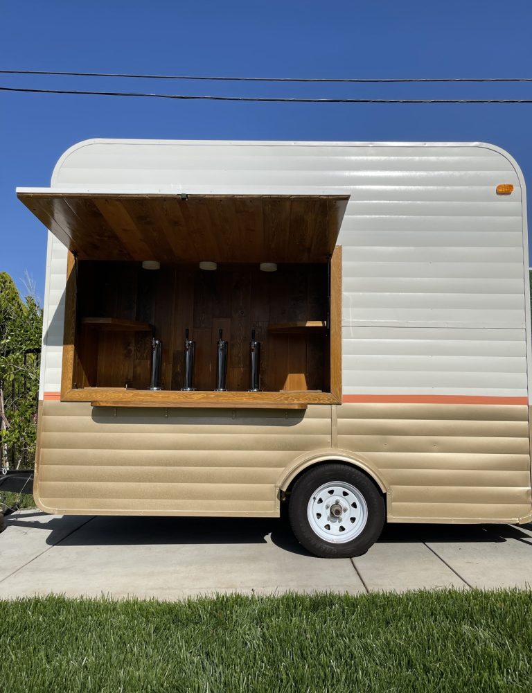 A wide shot of a beer tap trailer from the side. The concession window is open. The interior has wooden planks, shelving on either side, and four taps at the center. The exterior of the trailer is white, with the lower half brown. A thin, orange strip borders the two.