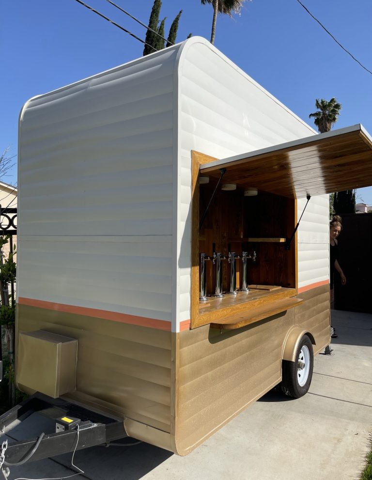 A wide shot of the beer tap trailer. The view is from the front, and slightly to the side, revealing the side of the trailer. The concession window is open. The interior has wooden planks, shelving on either side, and four taps at the center. The exterior of the trailer is white, with the lower half brown. A thin, orange strip borders the two.
