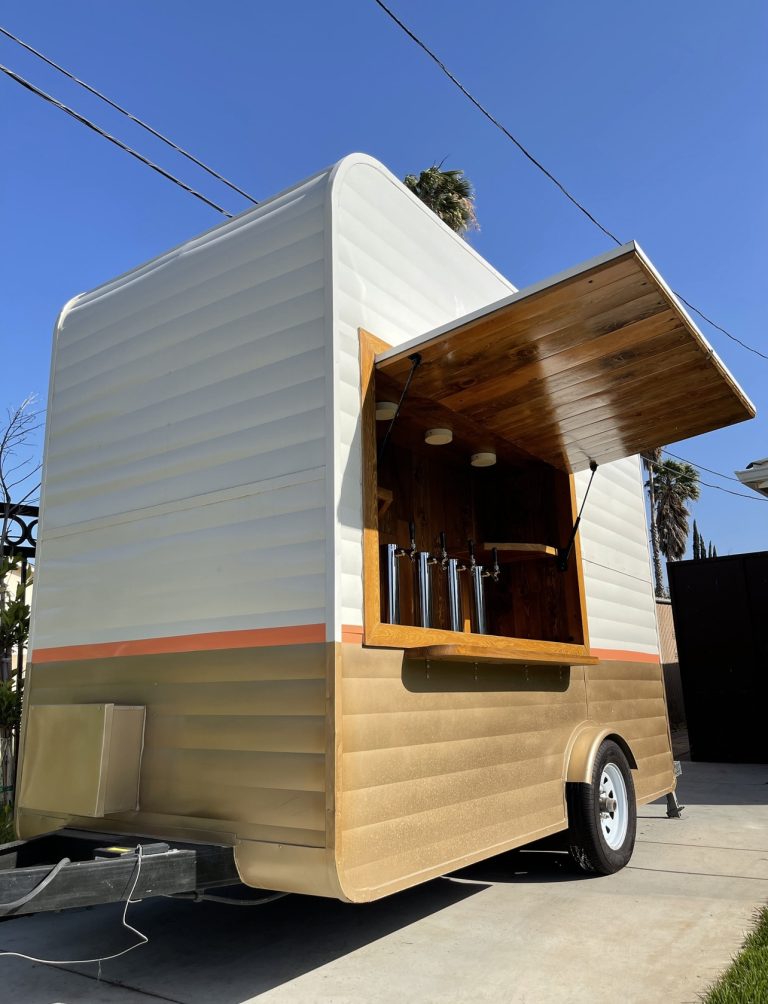A wide shot of the beer tap trailer. The view is from the front, and slightly to the side, revealing the side of the trailer. The image is taken at a lower angle, looking up at the trailer. The concession window is open. The interior has wooden planks, shelving on either side, and four taps at the center. The exterior of the trailer is white, with the lower half brown. A thin, orange strip borders the two.