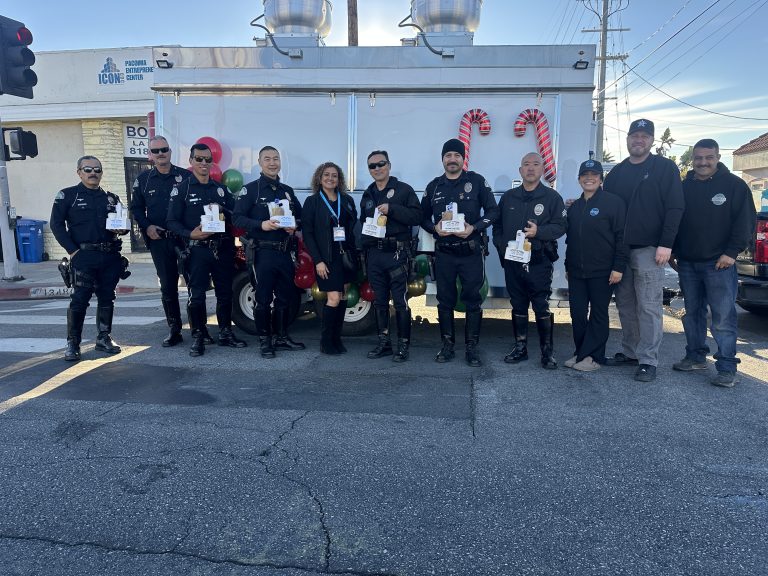 A wide shot with the The Food Truck Group's members standing together with the Los Angeles police for a picture. The police have carriers of coffee and tamales. Behind the group is a white food trailer with balloons and candy canes. Above them is a clear sky.