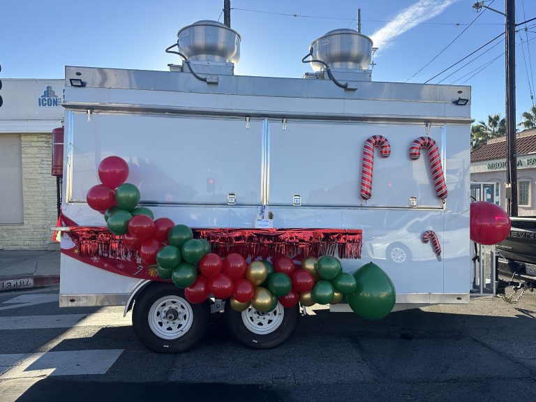 A wide shot of a food trailer. Balloons of red, green, and gold are on the left side. On the right are three candy canes, two big ones and one small one below them.