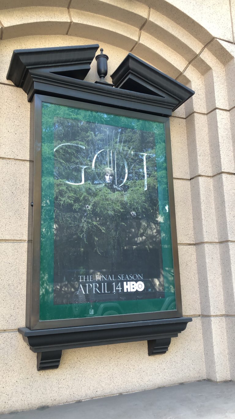 Vertical shot of a theater poster display case. The poster has the letters ‘GOT’ in the center for Game of Thrones, with a green border outline. The bottom has the text ‘The Final Season April 14 HBO’. The poster features the season’s antagonist seated on the throne.