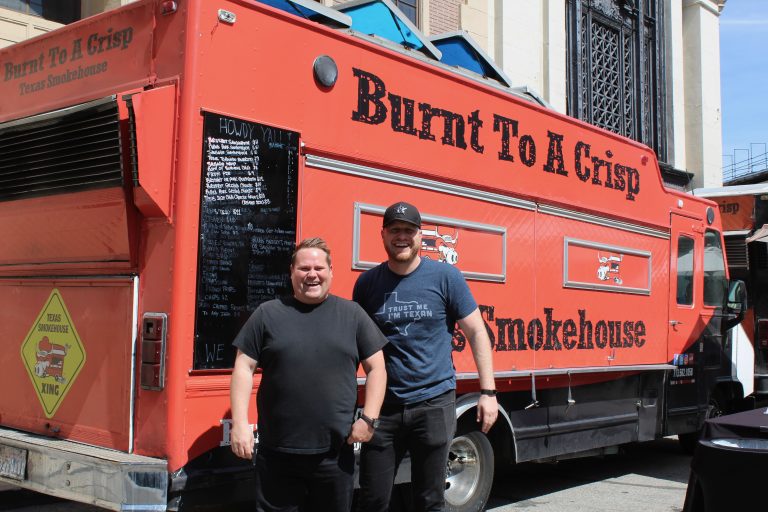 A wide shot of Burnt To A Crisp’s food truck. The service side is visible. To the left of it is a black board with the menu written in white text. The food truck is red, and faces away from the camera. Before the food truck are the owners. The men stand together, smiling at the camera. Behind the food truck is a building at Warner Brothers. It is morning.