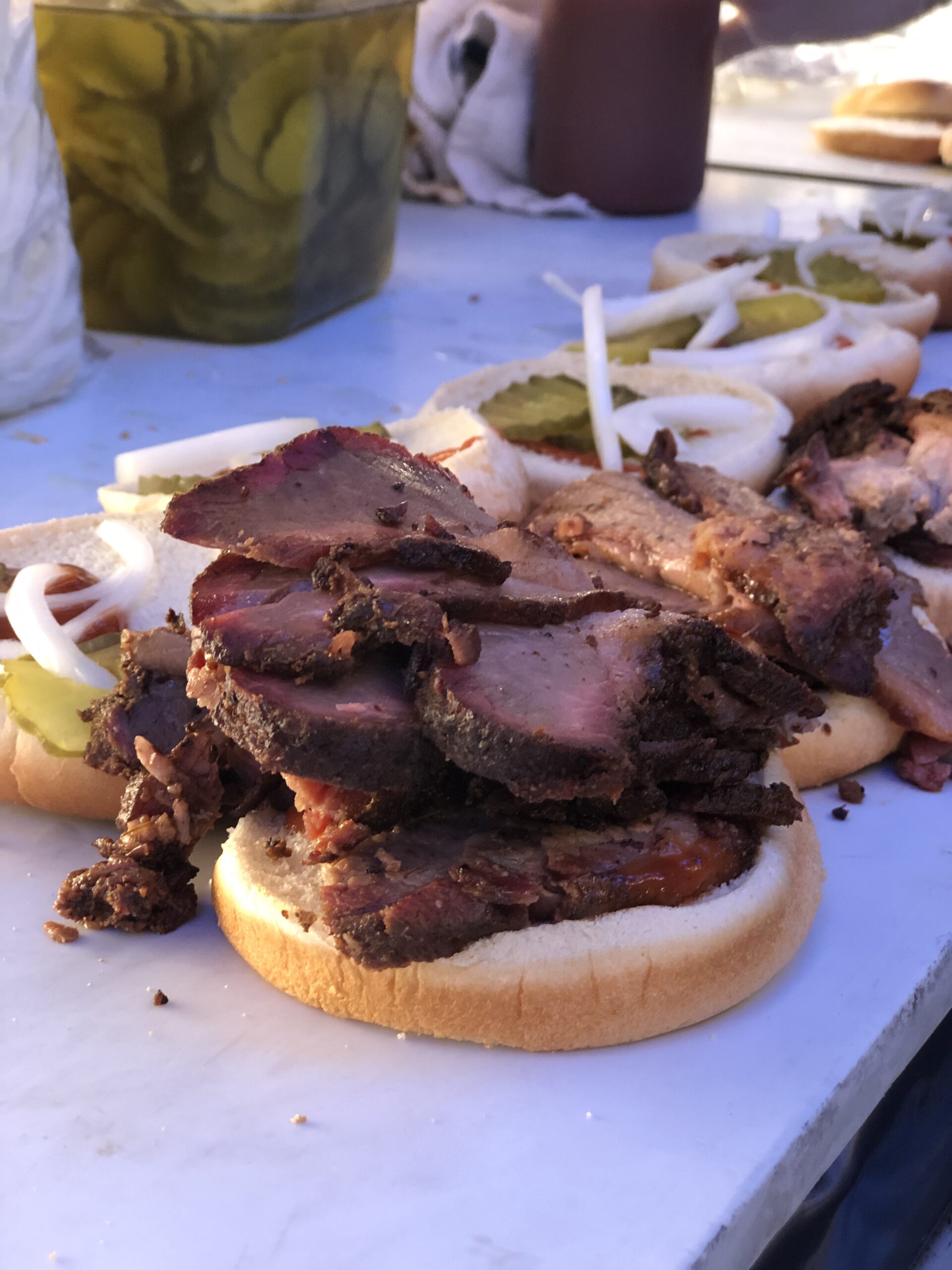 A vertical close-up shot of a brisket hamburger on a counter. The top bun is off, with onion, pickle slices, and ketchup covering it. The bottom bun has brisket stacked over it. A row of similar hamburgers lead into the distance of the shot.