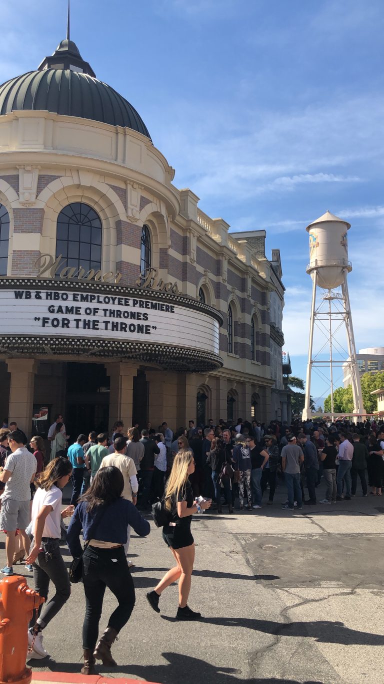 A vertical shot of the movie theatre Warner Bros. The marquee reads, ‘WB & HBO EMPLOYEE PREMIERE GAME OF THRONES “FOR THE THRONE”’. A large crowd stands at the entrance in long lines. In the distance is the Warner Bros water tower. It is morning.