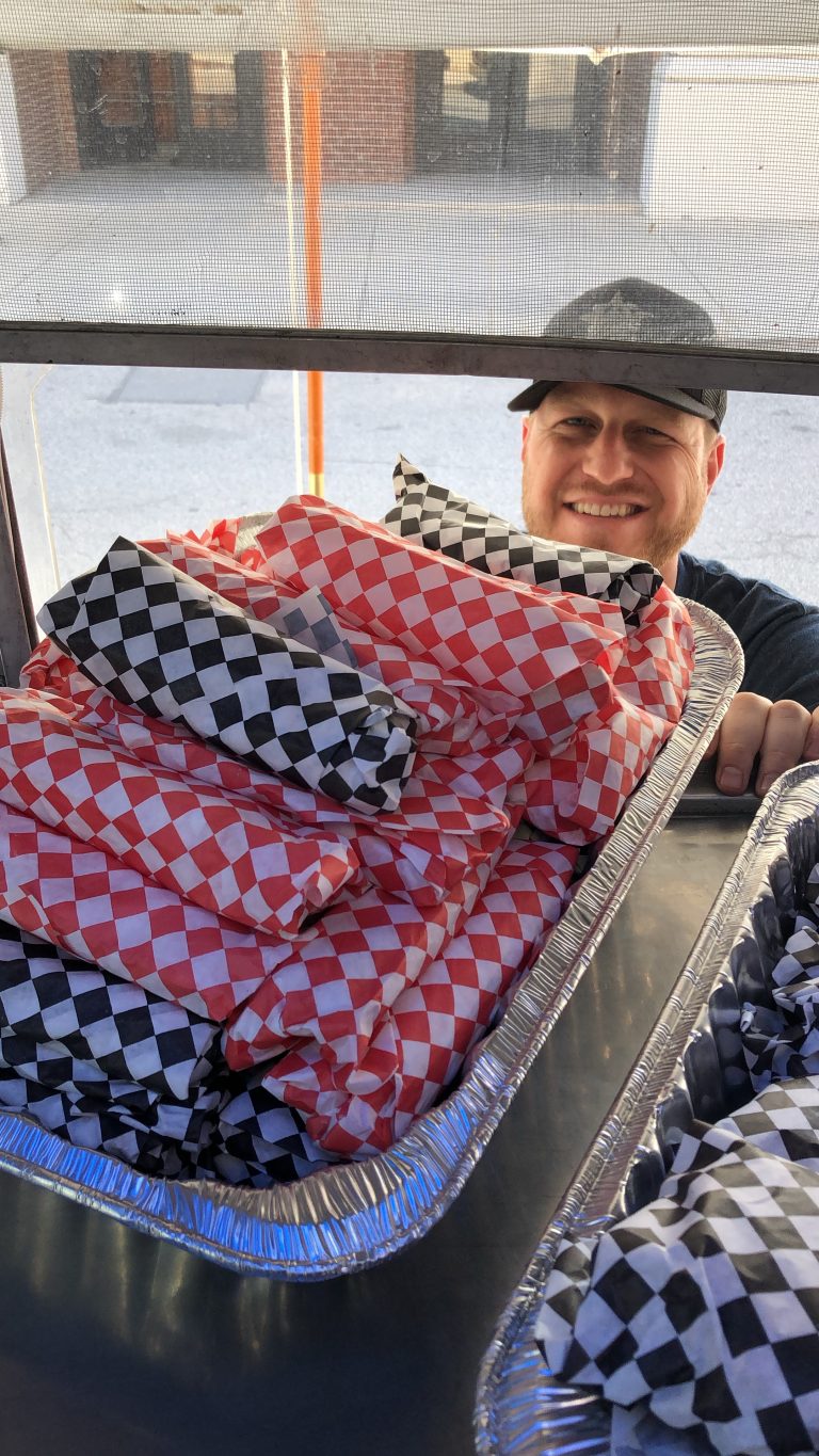 Close-up vertical shot of Burnt To A Crisp’s food truck interior. In the foreground is a metal countertop, with two trays full with hamburgers in checkered wrapping paper. Just outside of the open service side window is the owner, smiling. The background shows the floor, with a small portion of a building at the very back. It is morning.