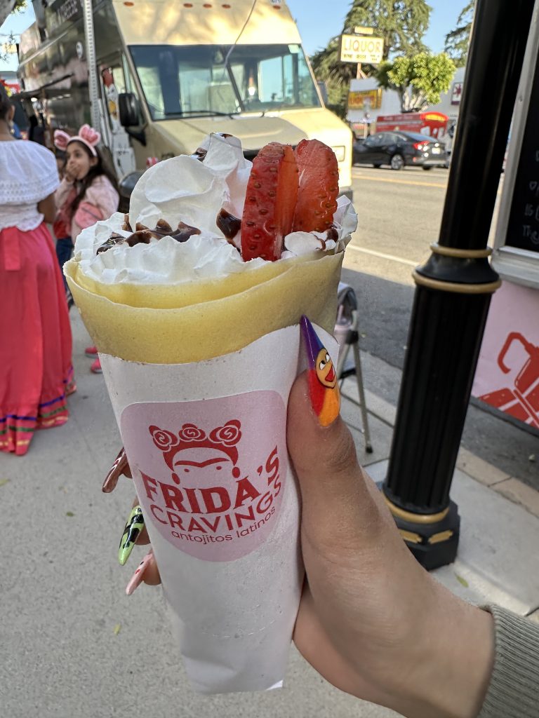 A close shot of a woman’s hand holding a crepe folded like a cone, containing whipped cream with chocolate drizzle on top. Around the crepes is a paper wrapper. On the center of the wrapper is a pink, circular sticker with the words, ‘Frida’s Cravings antojitos latinos’ and a logo of the company on top. Behind the crepe is the sidewalk, with another food truck behind and some people.