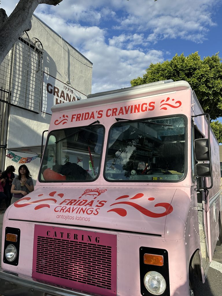Vertical shot of Frida’s Cravings cabin. Above the windshield is Frida’s Cravings name. On the hood is the business’s name and logo. To the left of the food truck are customers standing at the side. The blue sky is streaked in clouds.