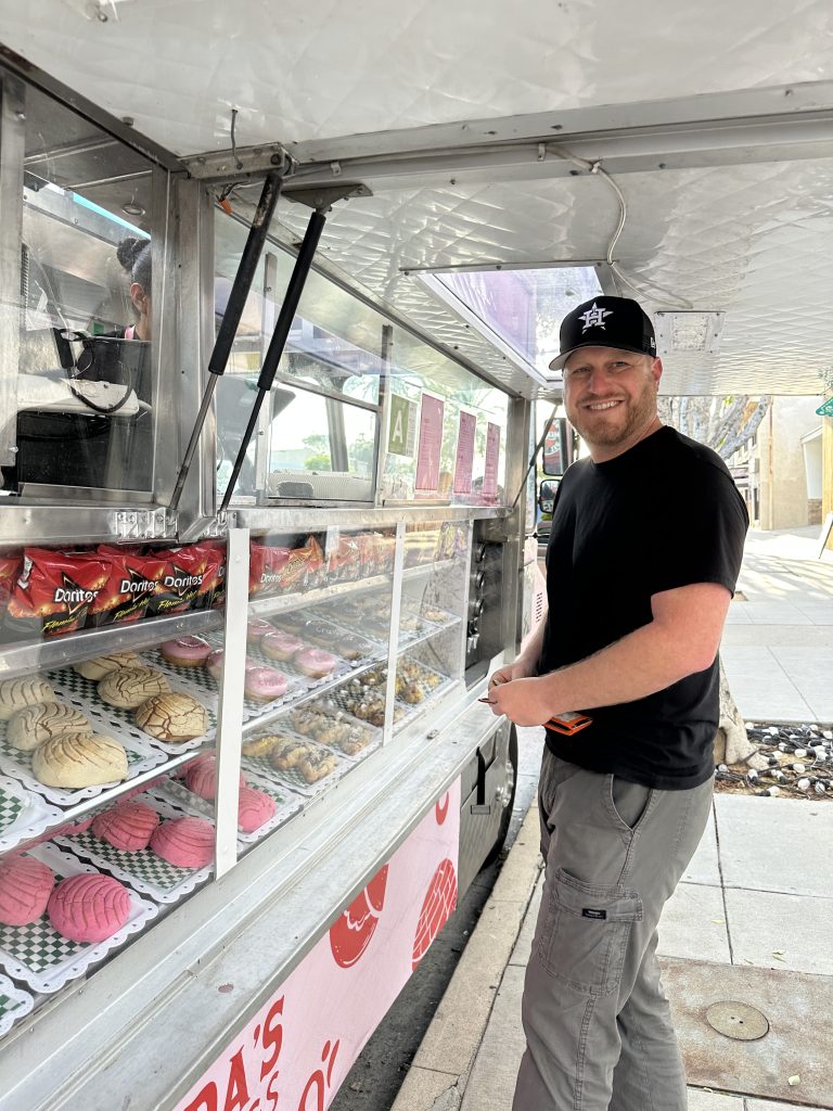 Vertical shot of Frida’s Cravings and a customer. The view is directly beside the food truck looking down the street. To the left is the food truck, service window open to show pastries behind the glass. A service window attendant stands at the ready. To the right is the male customer, smiling at the camera.