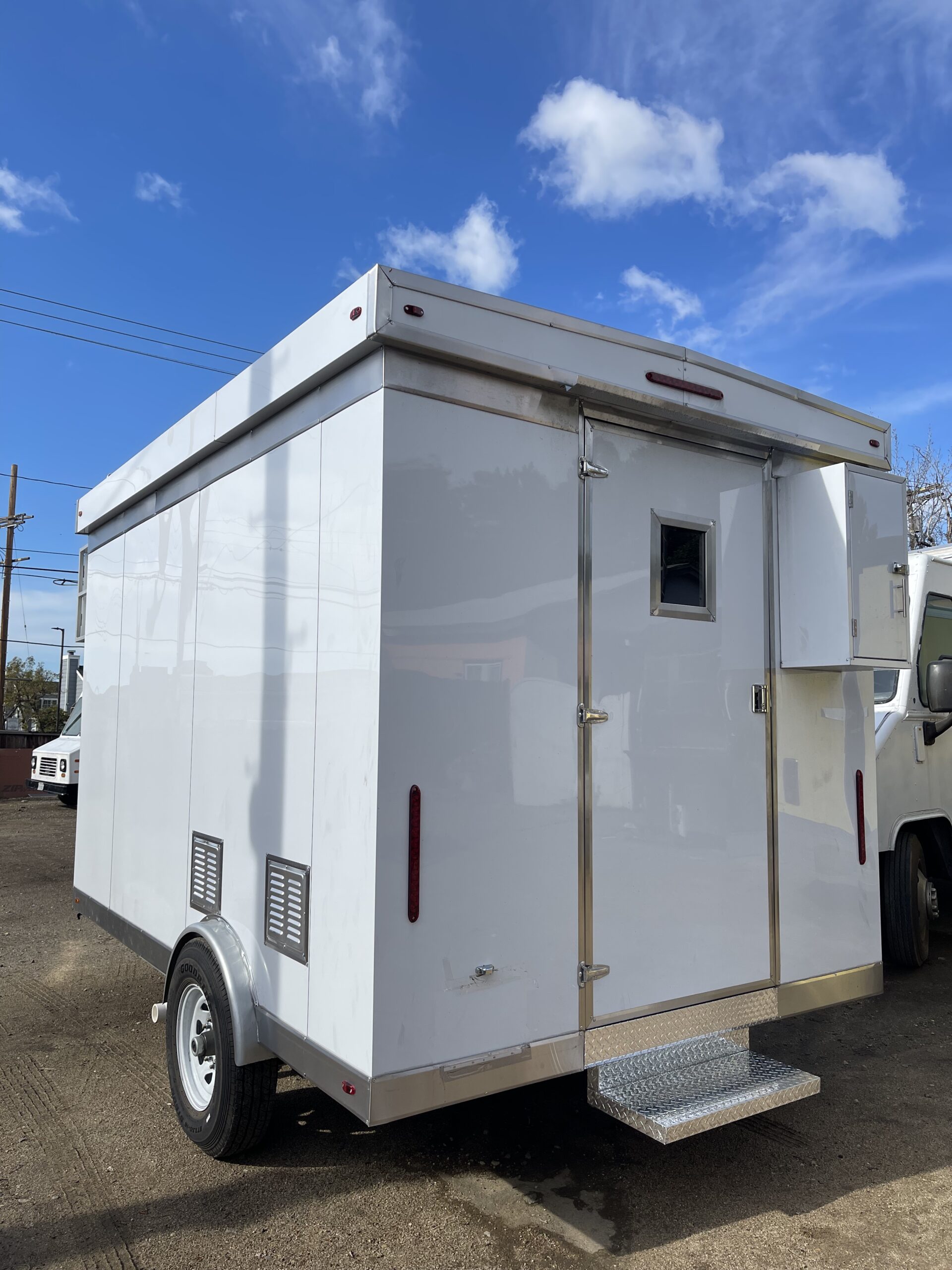 A wide shot of a white food trailer with two wheels. The door to the trailer is visible, with a steel step attached to the trailer. The trailer is on dirt below a cloudy sky.