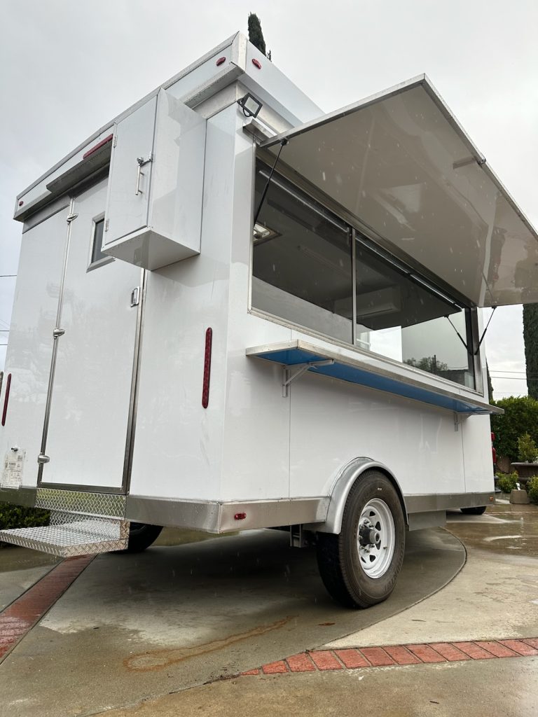 A wide shot of a white food trailer on two wheels, viewed from the side. The service side window is open. Facing closer to the viewer is the entrance to the food trailer, with a steel step attached to the trailer.