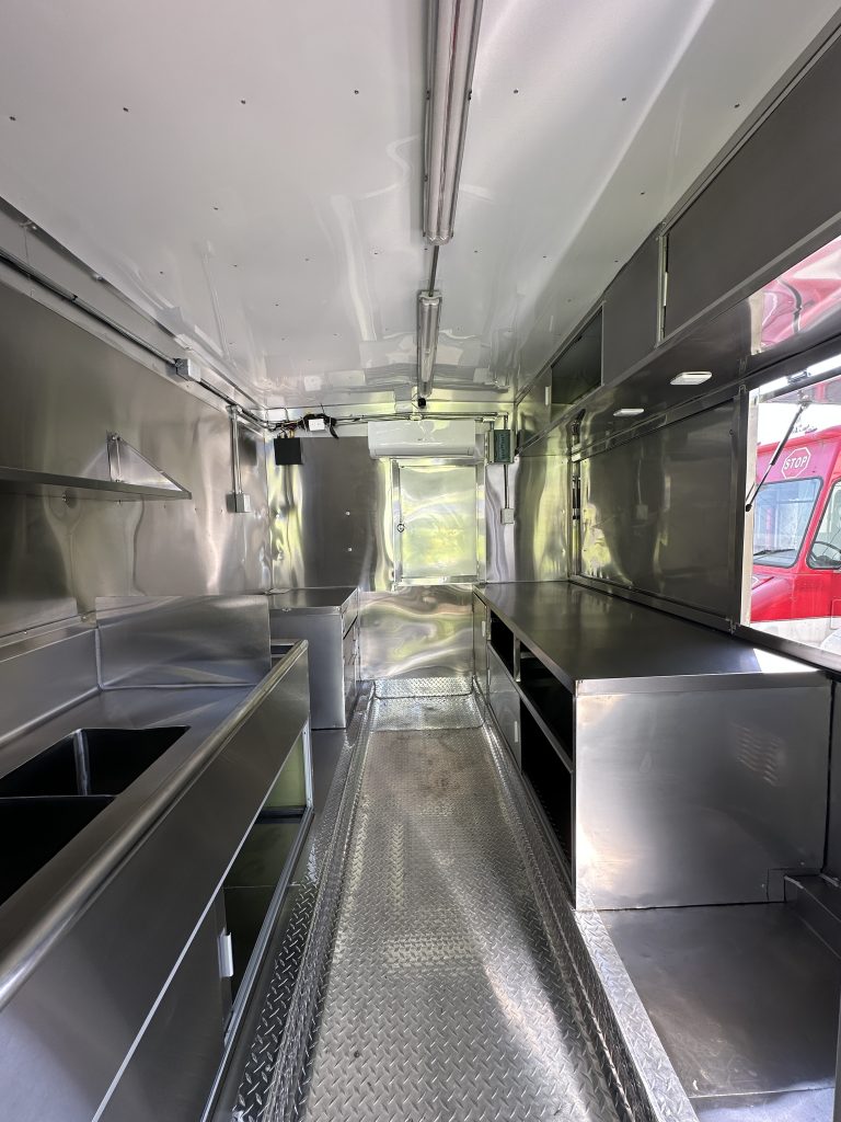 An interior shot of a food trailer. The interior is made of stainless steel with shelving and a compartment sink. The right side has the service side window. The photo was taken from the entrance looking ahead into the trailer.