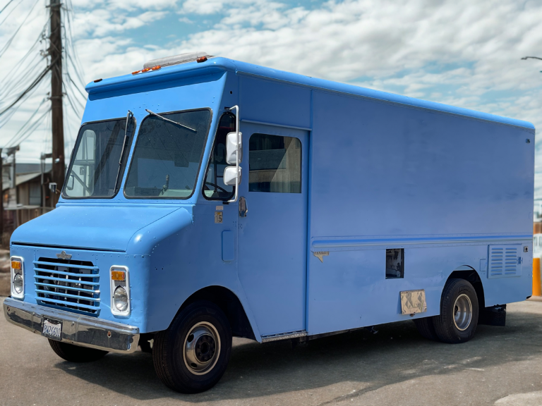 A wide shot of a blue food truck from the driver's side view. The food truck is on a gravel road.