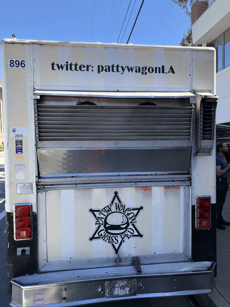 A portrait shot of the back of The Patty Wagon. At the bottom half is the logo of the food truck, which features a six pointed star and a hamburger in the center, and the text ‘Patty Wagon Grass Fed’ surrounding the hamburger. On the upper half is the text ‘twitter: pattywagonLA’.