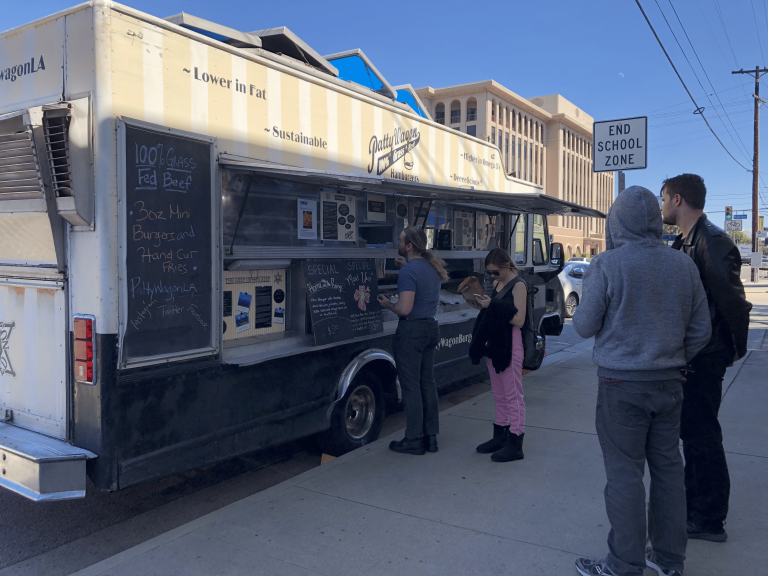 A wide shot of The Patty Wagon. The upper half is striped in yellow and white, the bottom half black. The picture is taken from the side of the food truck, with the service side window open. Customers are lined beside the food truck. The food truck is parked against the sidewalk.