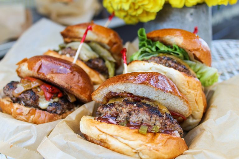 Close up shot of four hamburgers from The Patty Wagon. The burgers are on top of paper trays with wax paper beneath them. Each hamburger has a toothpick on the bun. Each also has different toppings.