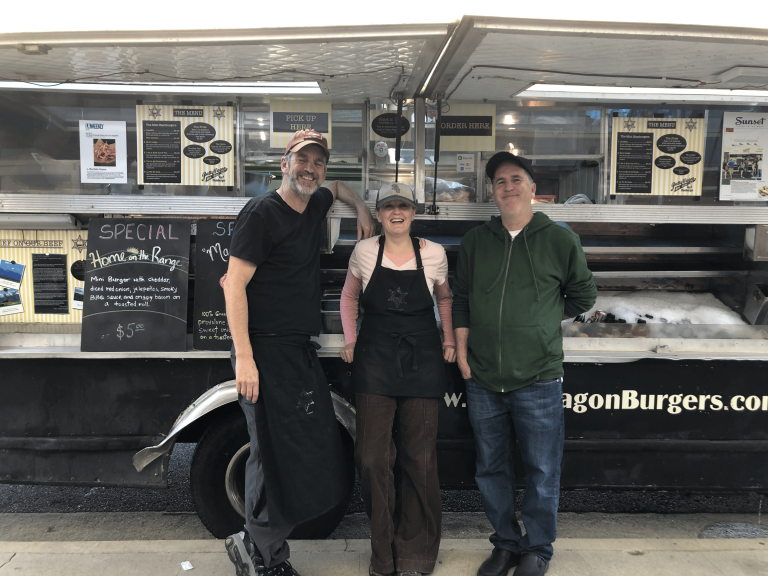 wide shot of The Patty Wagon from the open side view window. In front are the three owners, standing close together and smiling at the camera. To the left of them is a black board with the current special. To the right in the image is the drinks container, with ice keeping them cool.