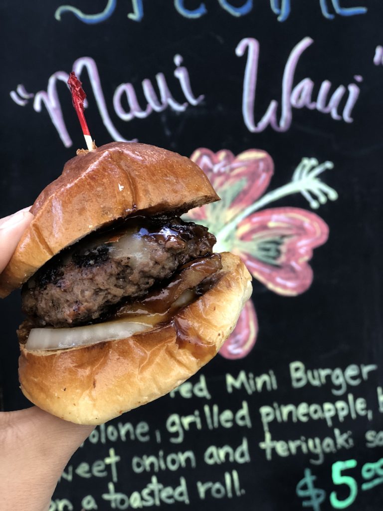 A portrait shot of a hamburger held in a hand. The hamburger is close to the viewer. Behind the hamburger is a blackboard with a flower drawn just to the right, slightly behind the burger. On the top is the name of the dish being served, and at the bottom the text lists the ingredients.