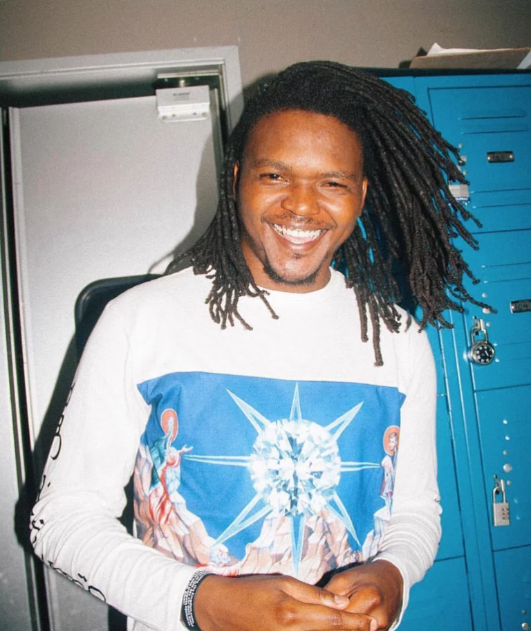 Portrait shot of Young Nudy from the waist up. He is smiling. His body is leaned slightly to the side, with his hands close together. He wears a white shirt with a large, blue image over the chest. He stands beside a few blue lockers.