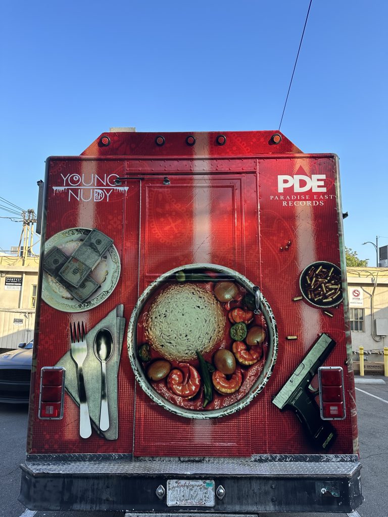 Portrait shot from the back of Young Nudy’s food truck. The wrap is red. In the center is a dish with shrimp, sauce, and mushrooms. The wrap also features a gun with bullets on the right, two stacks of cash on the left of the dish, and two utensils atop a napkin. On the upper left of the wrap is the text ‘Young Nudy’, and on the far right, ‘PDE Paradise East Records’.