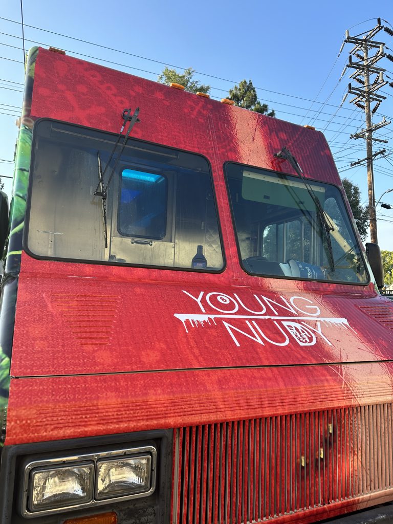 A portrait shot of the cabin of the food truck. The cabin has a red wrap over it. The hood of the food truck has the text “Young Nudy” in the center, with the first text above the second. Between them is a horizontal line.