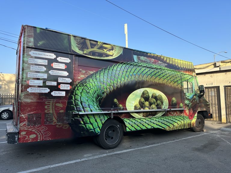A wide shot from the service side of the food truck. The wrap is red with circular patterns. In the center of the food truck’s side is a plate of brussel sprouts. Coiled around it is a large, emerald snake, its head located on the cabin door.