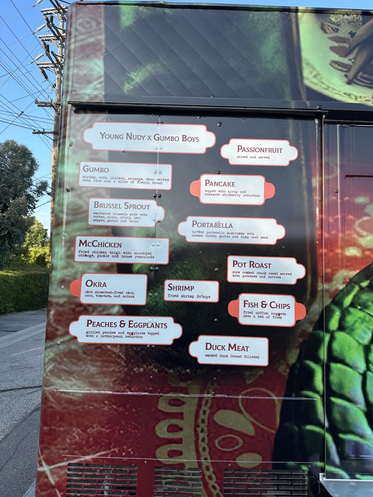 A portrait shot from the service side of the food truck. The photo is zoomed in close to the side end of the truck, where the menu is visible. The wrap of the food truck is red, but darker where the menu is. The menu is made on the wrap, with a white border and red outline to make them stand out. There are twelve menu options featured. Above the text is the words, “Young Nudy x Gumbo Boys.”