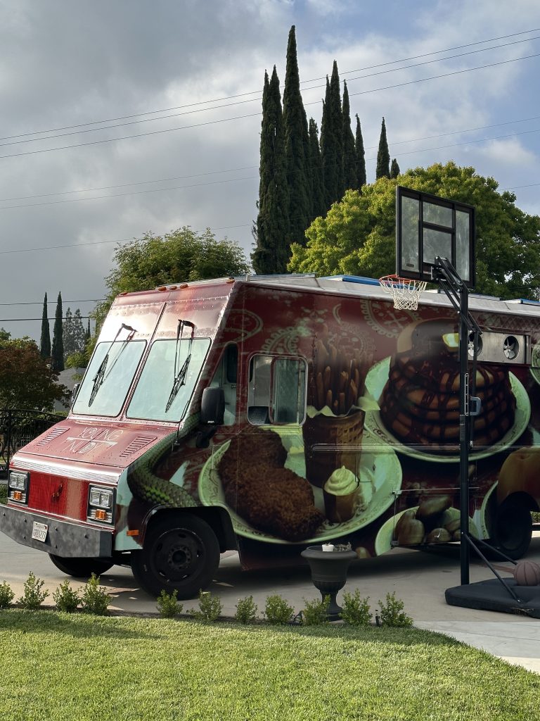 wide shot of the food truck. It is on a driveway, behind an outside basketball hoop. The side of the food truck is visible, with a red wrap. The wrap features plates of food directly from the menu, including a plate of pancakes. Along the sides of the wrap is the emerald snake image moving around the plates.