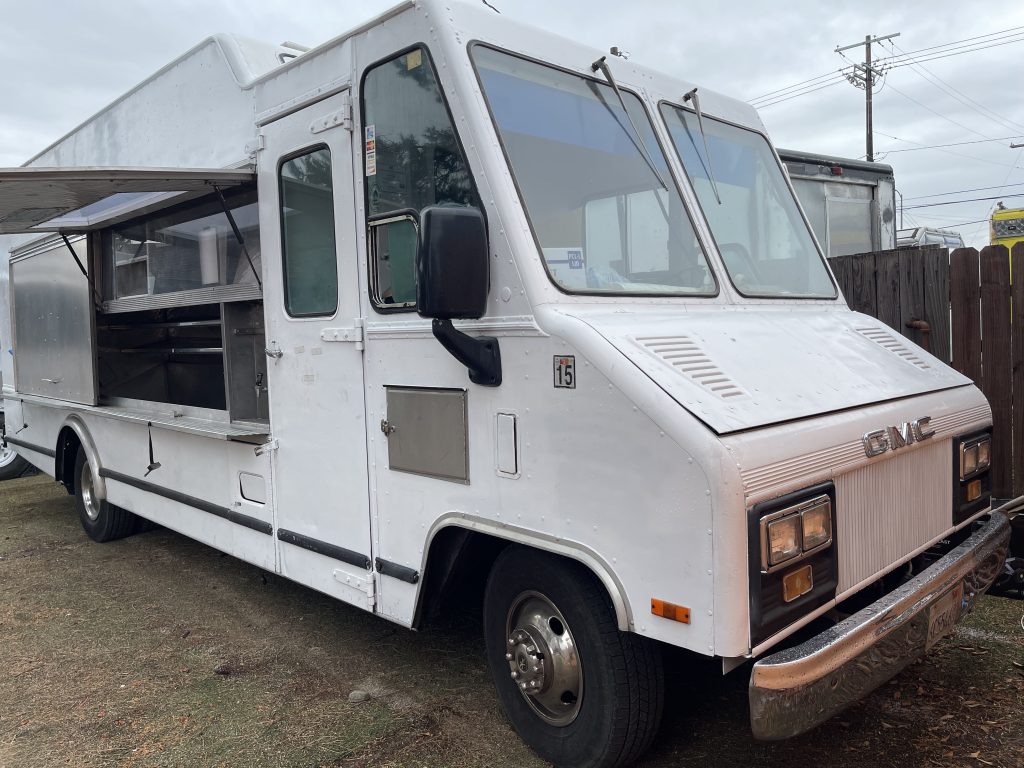 A wide shot of a white food truck. It is parked on the dirt.