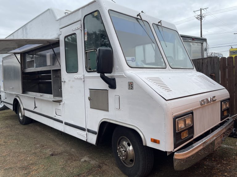 A wide shot of a white food truck. It is parked on the dirt.