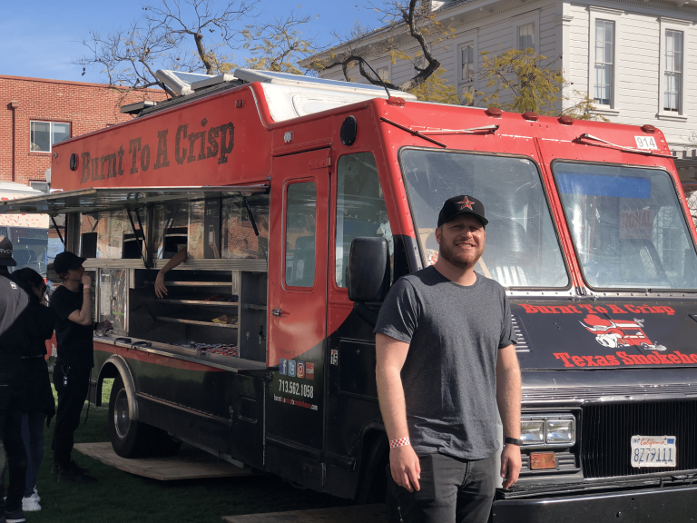 A wide shot of the Burnt To A Crisp food truck. The food truck is red and black. The owner, a man, is stood in front of the food truck.